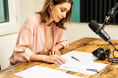 Merina الدين in een lichte podcaststudio terwijl zij een interviewscript voorbereidt aan een houten tafel